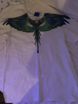 Vit t-shirt med färgglada vingar - Säljer en vit t-shirt med ett unikt tryck av gröna och blå vingar över axlarna och bröstet. T-shirten har rund hals och korta ärmar. Perfekt för dig som vill sticka ut med en cool och konstnärlig design.
