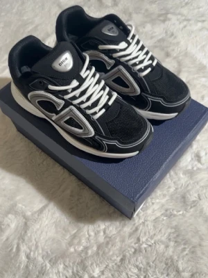 Svarta Dior B30 sneakers - Snygga svarta Dior B30 sneakers med vita och grå detaljer. Skorna har en sportig design med mesh och läderpaneler, samt tydliga Dior-loggor på sidan och plösen. Vita snören och en chunky sula ger en modern vibe. Perfekta för dig som vill sticka ut med exklusiva sneakers.