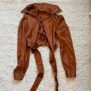 Brun satinskjorta med knytband - Snygg brun blus i satin med långa ärmar och bred krage. Framtill finns dekorativa knytband och rynkade detaljer som ger en trendig look. Perfekt för dig som gillar statement-plagg och vill sticka ut lite extra.