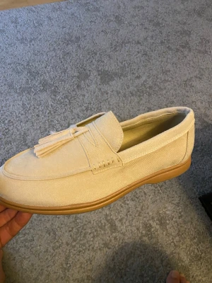 Beige loafers med tofsar - Snygga beige loafers i mockaimitation med dekorativa tofsar framtill och ljusbrun sula. Skorna har en klassisk siluett och passar perfekt till en avslappnad men stilren look. Materialet ger en mjuk känsla och de är lätta att matcha med olika outfits.