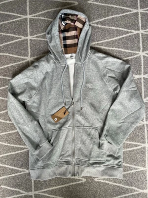 Grå zip hoodie från Burberry - Snygg grå hoodie med dragkedja från Burberry. Klassisk rutmönstrad insida i huvan och broderad logga på bröstet. Tillverkad i mjuk bomullsblandning, har två fickor fram och justerbar dragsko i huvan. Perfekt för en chill och stilren look.