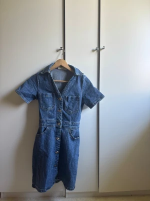 Blå jeansklänning med knappar - Snygg blå jeansklänning med kort ärm, knappar framtill och fickor både upptill och nertill. Klänningen har klassisk skjortkrage och är tillverkad i robust denim. Fligurnära och kort i modellen. Perfekt för dig som gillar en avslappnad men trendig stil. Använd sparsamt utan slitage.