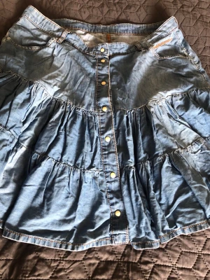 Blå volangkjol i denim från Peak Performance - Säljer en blå kort volangkjol i bommull från Peak Performance i denim med tryckknappar framtill och broderad logga på fickan. Kjolen har flera lager med volanger och coola detaljer som kontrastsömmar. Perfekt för dig som gillar en avslappnad och trendig look.