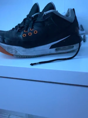 Svarta Nike Air Jordan sneakers - Säljer ett par svarta Nike Air Jordan sneakers med orange detaljer och vit sula. Skorna har synlig Air-enhet i hälen, lågt skaft och klassisk Jordan-logga på plösen. Ovandelen är i läder och textil, och sulan har orange undersida.