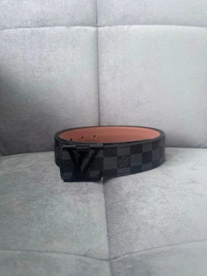 L Bälte Svart - Snyggt svart bälte från L med klassiskt Damier-mönster och L-logga på spännet. Bältet är gjort i läder med svart och grå rutigt yttre och ljusbrun insida. Perfekt för att lyfta din outfit med en exklusiv touch.