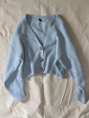 Ljusblå kofta från H&M Divided - Mysig ljusblå stickad kofta från H&M Divided med tre träfärgade knappar framtill. Koftan har v-ringning, ribbstickat mönster och är lite croppad i modellen. Perfekt att slänga på sig över en t-shirt eller linne.