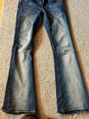 Blå bootcut jeans från Gina Tricot - Snygga blå bootcut jeans från Gina Tricot med röda kontrastsömmar och klassisk femficksdesign. Jeansen har en lätt tvättad look och dragkedjegylf. Perfekta för dig som gillar en retrostil med utsvängda ben.