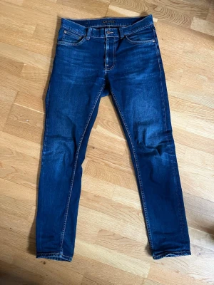Mörkblå Nudie jeans - Snygga mörkblå slim jeans från Nudie med klassisk femficksdesign och orangea sömmar. Litet hål i bakfickan (se bild) annars inga problem. Storlek: 31/32 Modell: Lean Dean