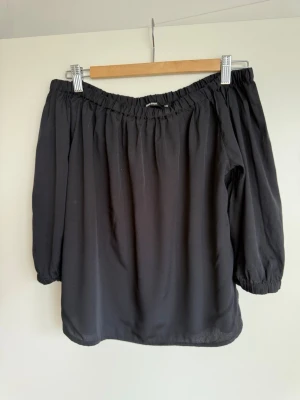 Svart offshoulder blus från Gina Tricot - Trendig svart offshoulder blus från Gina Tricot med trekvartsärmar och resår vid axlar och ärmslut. Blusen har en luftig passform och är tillverkad i ett lätt, vävt material som ger en snygg siluett. Perfekt att styla med jeans eller kjol.