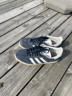 Adidas Gazelle blå och vita sneakers - Adidas Gazelle sneakers i blå mocka med vita läderdetaljer och de klassiska tre ränderna på sidorna. Låg modell med vit sula och vit hälkappa. Perfekt för dig som gillar retro och streetstyle.