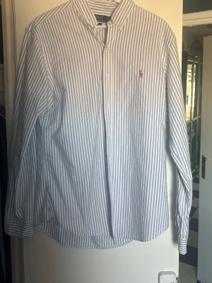 Randig skjorta från Polo Ralph Lauren - Klassisk vit skjorta med blå ränder från Polo Ralph Lauren. Slim fit-modell med knappar framtill och broderad logga på bröstet. Långärmad och tillverkad i bomull, perfekt för en stilren look.