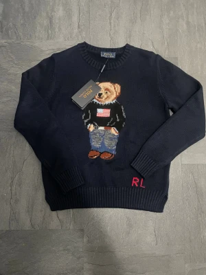 Mörkblå Polo Ralph Lauren björntröja - Mörkblå stickad tröja från Polo Ralph Lauren med ikonisk björn på bröstet, klädd i jeans och svart tröja med amerikansk flagga. RL-broderi vid nederkanten. Rund hals och ribbade muddar. Perfekt för dig som gillar klassisk preppy stil. Jag säljer den för att jag råkade köpa fel jag såg inte att den var för tjej .