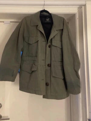 Militärgrön tygjacka från Zara - Snygg militärgrön tygjacka från Zara med fyra stora fickor framtill och bruna knappar. Jackan har en rak passform, klassisk krage och är tillverkad i ett mjukt bomullsliknande material. Perfekt för lager-på-lager och streetstyle.