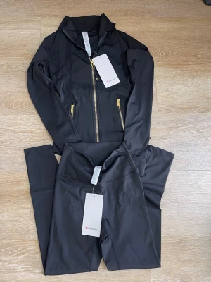 Svart träningsset från Lululemon - Snyggt svart träningsset från Lululemon med jacka och tights. Jackan har dragkedja i guld och hög krage, medan tightsen har hög midja. Perfekt för gymmet eller löprundan. Materialet är stretchigt och följsamt för maximal rörelsefrihet.