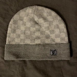 Grå mössa - Snygg grå mössa med diskret rutmönster och uppvikt kant. logga framtill på kanten. Tillverkad i mjukt och bekvämt material som håller dig varm under kalla dagar.