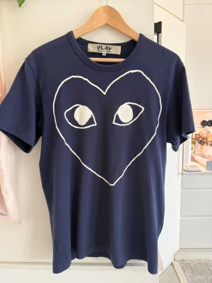 Mörkblå Comme des Garçons PLAY t-shirt - Snygg mörkblå t-shirt från Comme des Garçons PLAY med det ikoniska hjärtmotivet och ögon i vitt på bröstet. Klassisk rund hals och korta ärmar. Tillverkad i mjuk bomull som känns skön mot huden. Perfekt för en trendig och avslappnad stil.