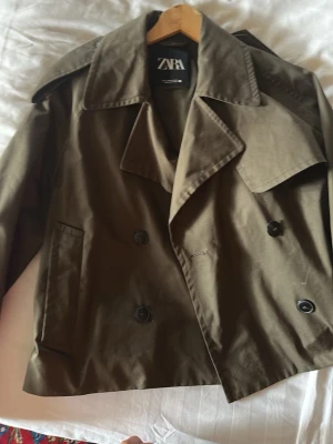 Olivgrön trenchcoat från Zara - Kort trenchcoat i olivgrönt från Zara med dubbelknäppning, breda slag och axelklaffar. Jackan har två snedställda fickor framtill och klassiska detaljer som stormflik bak. Perfekt för en trendig och stilren look.