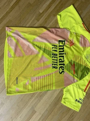 Arsenal målvaktströja neon Adidas - Cool Arsenal målvaktströja från Adidas i neon-gult med rosa och gröna detaljer samt svarta tryck. Tröjan har korta ärmar, V-ringning och klubbmärke på bröstet. Materialet är lätt och andas, perfekt för fotbollsträning eller match.