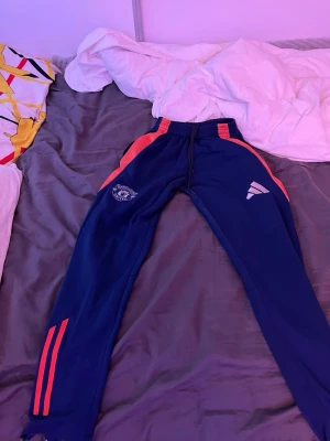 Blå Manchester united tränings byxor  - Snygga blå mjukisbyxor från Adidas med röda ränder och detaljer längs sidorna. Byxorna har resår i midjan med dragsko och tryckt logga på ena benet. Perfekta för chill eller träning, med smal passform och mjukt material.PRISET KAN DISKUTERAS!