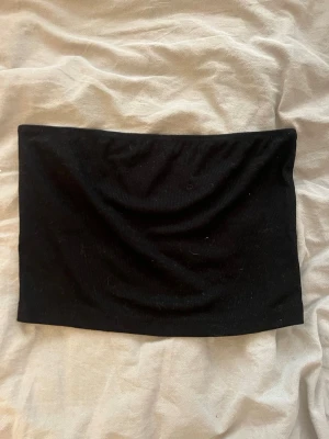 Svart bandeau tubtopp - En enkel och stilren svart bandeau tubtopp i ribbat material. Perfekt att styla med jeans eller kjol för en clean look. Toppen är ärmlös och har en tight passform som sitter snyggt på kroppen.