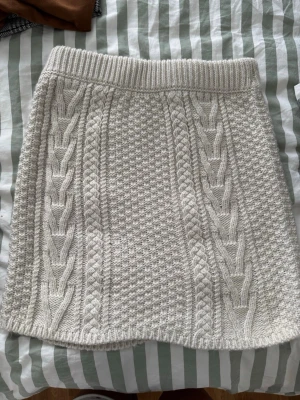 Vit stickad kortkjol med flätmönster - Superfin vit stickad kortkjol med snygga flätade detaljer och bred resår i midjan. Kjolen har ett klassiskt mönster och är perfekt för dig som gillar en mysig och trendig stil.