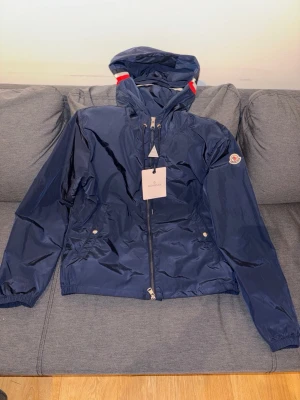 Mörkblå Moncler vindjacka med huva - Snygg mörkblå vindjacka från Moncler med huva och dragkedja framtill. Jackan har vita och röda detaljer på huvan, två fickor med tryckknappar och Moncler-logga på ärmen. Tillverkad i glansigt, lättviktigt material som står emot vind.