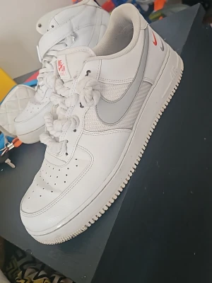 Nike Air Force 1 vita sneakers - Klassiska Nike Air Force 1 sneakers i vitt läder med grå swoosh och röd Nike-logga på hälen. Skorna har perforerad tå, tjocka vita snören och låg profil. Perfekta för dig som gillar en clean och tidlös streetstil.