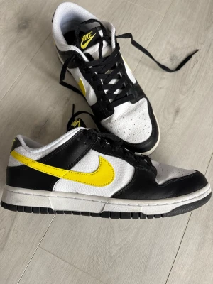 Nike Dunk Low svart/vit/gul - Nike Dunk Low sneakers i svart och vitt läder med gula swoosh-detaljer. Klassisk siluett med svart snörning och perforerad tåbox för extra ventilation. Tydlig streetstil och ikonisk design som sticker ut.