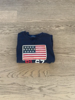 Blå sweatshirt från Polo Ralph Lauren - Mörkblå sweatshirt med amerikansk flagga och texten RL67 på bröstet. Klassisk rund halsringning och långa ärmar. Perfekt för dig som gillar streetwear och vill ha en stilren men ändå statement tröja.