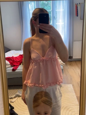 Ljusrosa volangtop med smala axelband - Supersöt ljusrosa topp med volangdetaljer och smala axelband. Toppen har v-ringning och är ärmlös, perfekt för varma dagar. Materialet känns lätt och luftigt, och modellen är loose för en avslappnad vibe.