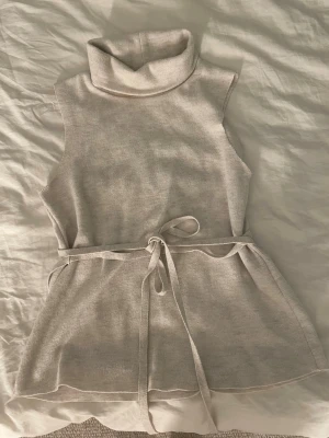 Beige ärmlös stickad polotröja - Säljer en beige stickad polotröja utan ärmar med knytskärp i midjan. Tröjan har hög krage och är gjord i mjukt material som ger en avslappnad och trendig look. Perfekt att styla över en långärmad topp eller skjorta.