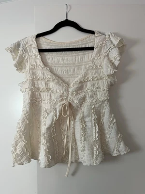 Söt volangblus med knyt i gul - Romantisk blus i ljusgul med volanger, spetsdetaljer och knyt framtill. Kortärmad modell med v-ringning och luftig passform. Perfekt för dig som gillar en feminin och bohemisk stil.