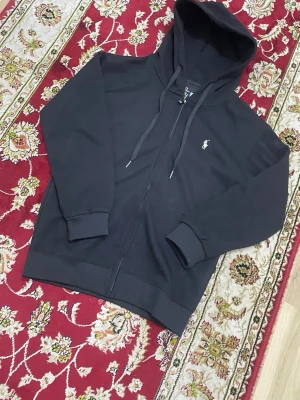 Svart hoodie från Polo Ralph Lauren - Svart hoodie med dragkedja från Polo Ralph Lauren. Klassisk design med huva och dragsnören, samt den ikoniska vita polospelaren broderad på bröstet. Mjuk bomullskvalitet och ribbade muddar vid ärmslut och nederkant. Perfekt för en chill och stilren look.