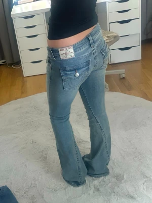 Ljusblå bootcut jeans True Religion - Säljer ett par ljusblå bootcut jeans från True Religion i modellen Joey med klassiska sömmar och silverfärgade knappar på bakfickorna. Jeansen har låg midja och är tillverkade i denim med lätt stretch. Det finns missfärgnings fläckar på nedre benen men är inte mycket synliga.