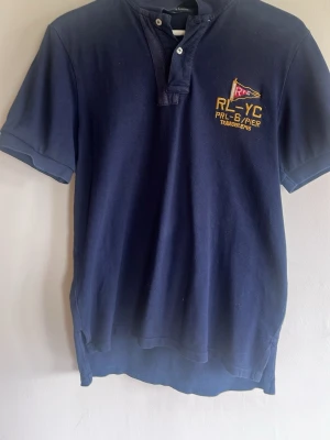 Marinblå pikétröja från Ralph Lauren - Marinblå pikétröja från Ralph Lauren med korta ärmar och tryck både fram och bak. Tröjan har knappslå vid halsen och broderad logga på bröstet. Perfekt för en sportig och avslappnad stil.