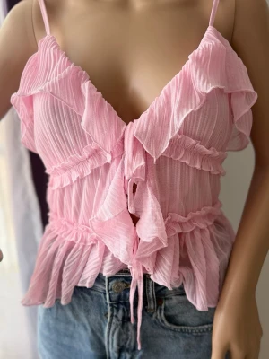 Rosa volangig linnetopp med knyt - Supersöt rosa linnetopp med volanger och knyt framtill. Tunt, lätt material med smala axelband och v-ringning. Perfekt för en somrig look och passar snyggt till jeans eller kjol.
