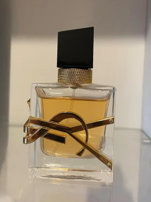 Yves Saint Laurent Libre Eau de Parfum - Säljer en lyxig parfym från Yves Saint Laurent, Libre Eau de Parfum. Flaskan är fyrkantig i genomskinligt glas med guldig YSL-logga och svart, stilren kork. Innehållet är bärnstensfärgat och flaskan har exklusiva guldiga detaljer runt halsen.