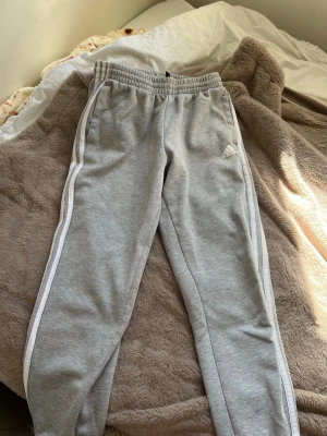 Gråa mjukisbyxor från Adidas - Snygga gråa mjukisbyxor från Adidas med vita ränder längs sidorna och klassisk logga på låret. Storlek 13-14 men är som storlek S