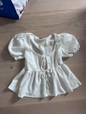 Vit blus med puffärm från Gina Tricot - Supersöt vit blus från Gina Tricot med puffiga korta ärmar och knytning framtill. Blusen har en lätt rynkad struktur och en feminin, utsvängd passform. Perfekt att styla med jeans eller kjol för en trendig look.