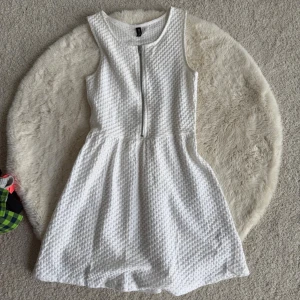 Vit ärmlös klänning från H&M Divided (XS) - Snygg vit ärmlös klänning från H&M Divided med strukturerat tyg och dragkedja framtill. Klänningen har rund halsringning och utställd kjol, perfekt för en fräsch och stilren look.