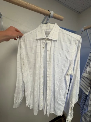 Vit rutig skjorta från Samuelsohn - Vit skjorta från Samuelsohn med diskret rutmönster i ljusblått. Klassisk krage och knappar framtill. Skjortan har lång ärm och är tillverkad i ett mjukt bomullsmaterial som känns skönt mot huden. Perfekt för dig som gillar stilrena och tidlösa plagg. Ordinarie pris: ca 1800-2500kr. Storlek M/L (15 1/2)