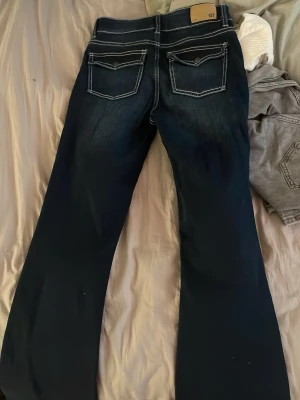 Bootcut jeans med kontrastsömmar - Mörkblå bootcut jeans med tydliga vita kontrastsömmar och fickor med lock bak. Jeansen har lätt slitning framtill och klassisk femficksdesign. Perfekta för dig som gillar en retro vibe och vill ha ett par jeans med lite extra attityd.