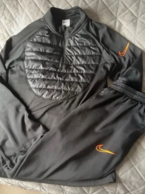 Svart Nike träningsset med orange detaljer - Snyggt svart träningsset från Nike med quiltad bröstpanel och halv dragkedja på tröjan. Både tröja och byxa har orange Nike-logga. Perfekt för träning eller chill. Materialet är mjukt och stretchigt för bästa komfort.