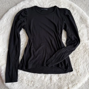 Svart långärmad topp från Zara - Snygg svart långärmad topp från Zara med rund halsringning och lätt puffade axlar. Tillverkad i mjukt och stretchigt material som sitter skönt mot kroppen. Perfekt att styla med jeans eller kjol för en trendig look.