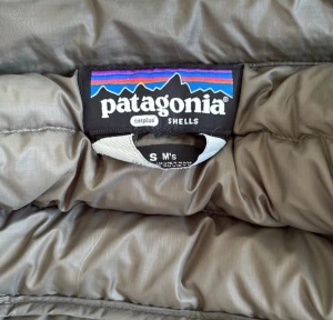 Grå/brun dunväst från Patagonia - Snygg grå dunväst från Patagonia med quiltad design och hög krage. Västen är lätt och smidig, perfekt för lager-på-lager och outdoor-looks. Märket syns tydligt på insidan och materialet är mjukt och följsamt. Den är använd några gånger men den har inte märken eller något som ser förstört ut på den. Lite osäker på material men jag tror de är Nylon, läs lappen på sista bilden. Storlek S/M
