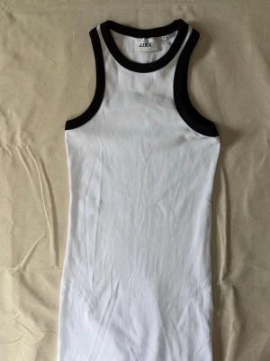 Vit ribbad kortklänning från JJXX - Säljer en vit ribbad kortklänning från JJXX med svarta kontrastkanter vid hals och ärmhål. Klänningen är ärmlös och har en kroppsnära passform. Aldrig använd.