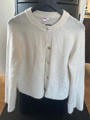 Vit kofta från Gina Tricot - Mysig vit kofta från Gina Tricot med rund halsringning och ribbade muddar. Koftan har knappar framtill och är tillverkad i ett mjukt, stickat material som passar perfekt för lager på lager. Enkel och stilren design som funkar till det mesta.