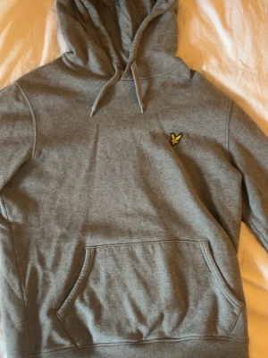 Grå hoodie från Lyle & Scott - Snygg grå hoodie från Lyle & Scott med klassisk gul logga på bröstet. Hoodien har huva med dragsko och en stor magficka framtill. Perfekt för en avslappnad och stilren look.