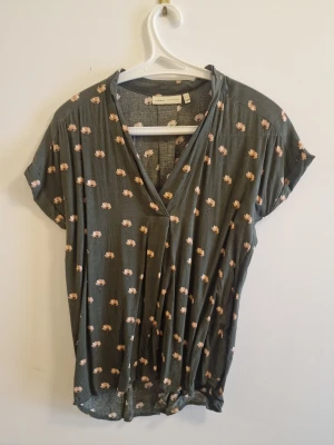 Grön blommig blus från InWear - Supersöt grågrön blus från InWear med små ljusrosa blommor och avslappnad passform. Djup V-ringning och korta ärmar ger en chill vibe. Perfekt att styla med jeans eller kjol. Materialet känns mjukt och svalt mot huden.