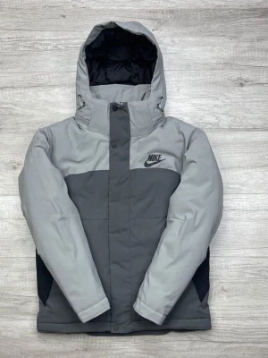 Grå pufferjacka från Nike - Snygg grå pufferjacka från Nike med två nyanser av grått och svart logga på bröstet. Jackan har huva, dragkedja med vindslå och svarta detaljer vid sidorna. Perfekt för kalla dagar och riktigt streetig stil.